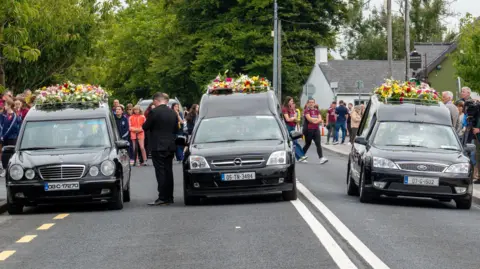 Victims of fatal shooting to be laid to rest in County Clare PA Media Three black hearses are lined up side-by-side on a road. Each has a flowers placed on top. There are a number of people standing behind the cars.