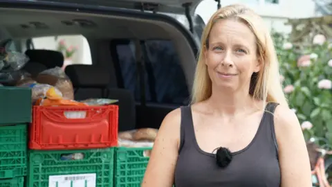 Rising Jersey food costs 'pushing families close to breadline' A woman with blonde hair and in a black tank top looking directly at the camera. Behind her is a car boot filled with crates of food.