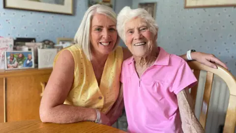 Parkrun record broken by 97-year-old Belfast runner Deirdre and Grace are sitting at a dining room table and both smiling at the camera. Deirdre is wearing a yellow top and Grace is wearing a pink t-shirt.