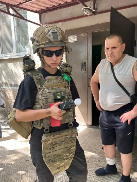 Inside Donetsk as residents flee attacks on Ukrainian region Putin wants to control Young woman Varia wearing khaki camouflage gear holding a device, standing next to a middle-aged man wearing a grey vest and blue trousers