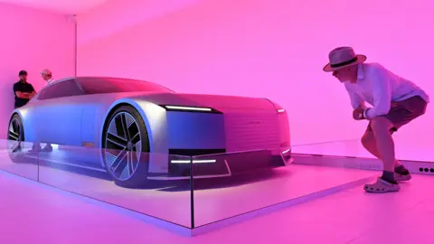PA Media A man wearing a wide-brimmed hat, white shirt, shorts and crocs crouches as he inspects a blue Jaguar 00 concept car, which is lit in hot pink. There are two people standing behind the car. 