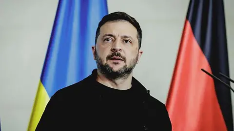 Trump shifts ceasefire stance and urges Ukraine to agree Russia peace deal Getty Images Ukrainian President Volodymyr Zelensky is wearing a black jacket over a black t-shirt, and looking above the camera. Behind him, there are two flags; one is blue and yellow, and the other is red and black.
