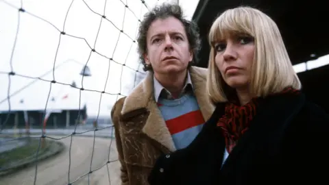 Ray Brooks, voice of Mr Benn, dies aged 86 Ray Brooks and Sharon Duce in character on the BBC programme Big Deal. Brooks wears a brown shearling jacket above a striped blue and red jumper. Duce has a short blonde bob and wears a large black jacket with a red and black patterned scarf.