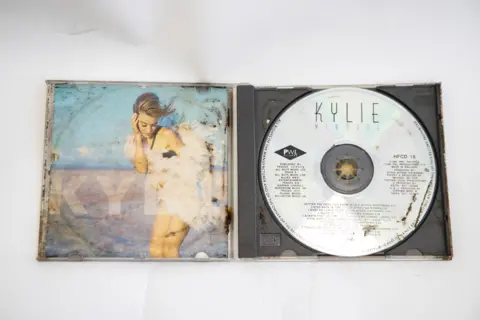 Princess Diana 90s time capsule contains Kylie CD and pocket TV Great Ormond Street Hospital for Children The CD of Kylie Minogue's album Rhythm of Love is open on a table. There is rust and dirt on different areas of the CD, which is metallic silver in colour.