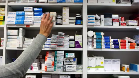 NHS to lose out on new drugs, pharma firm Novartis warns PA Media A person in a grey long-sleeve top reaching for a medication box on the top shelf in a pharmacy or medical storage area. The shelves are filled with neatly arranged boxes and containers of various medications, organised by type and brand.
