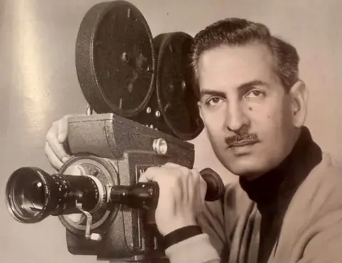 The WW2 veteran who moved Queen Camilla to tears Yavar Abbas A younger Yavar pictured beside his camera from his days as cameraman and film-maker. He has a moustache and looks straight into the camera with a serious expression.