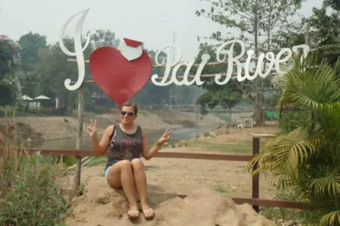 Man saw kaleidoscopic light before going blind Cheznye sits on a rock and poses in front of a sign for a photograph. A river can be seen behind her. She has light brown hair tied up and is wearing dark sunglasses.