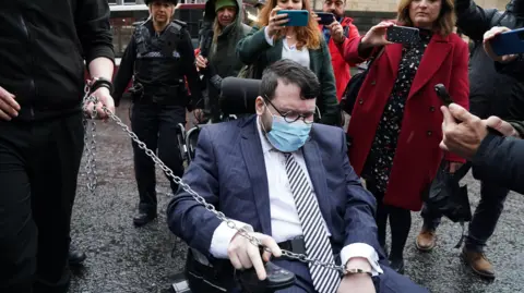 Fugitive Nicholas Rossi guilty of rape after extradition to US PA Media Nicholas Rossi outside court. He is in a wheelchair and is wearing a blue suit, white shirt, striped tie, glasses and an oxygen mask. People behind him are filming him on their phones and a long chain being held by a police officer links to his handcuffs.