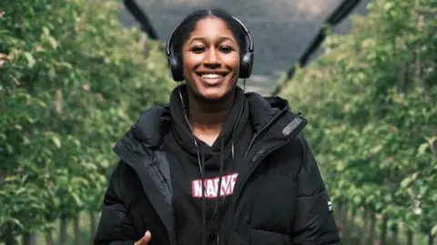I didn't go to uni Thaliqua Smith Thaliqua Smith stands smiling at the camera, with hedges either side of her. She has her black hair tied up and is wearing a pair of black headphones. She has a black Marvel hoodie on, with a black puffer jacket over the top.