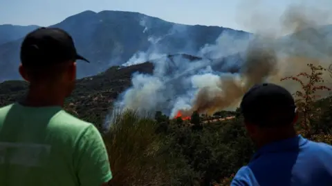 Wildfires rage across southern Europe as temperatures pass 40C Reuters Two people both wearing hats have their backs to the camera as they look on as smoke rises from a wildfire burning in the distance with white smoke and orange flames fanning