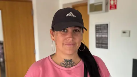 Free school uniform bank in Suffolk 'vital' say parents George King/BBC A head and shoulders image of Becky Mather. She is wearing a baseball cap and a pink jumper. She is looking into the camera. She has a large floral tattoo on her throat, and smaller tattoos down both sides of her face