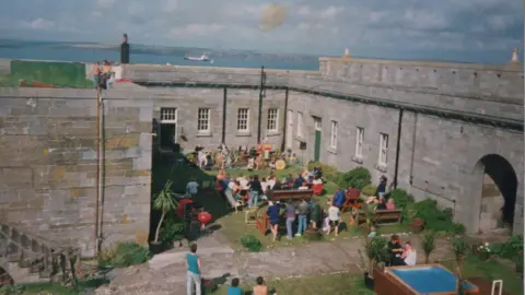 Pembrokeshire's Thorne Island fortress turned into £3m party space Mike Conner A group of people sit on benches in a courtyard, surrounded by large grey fortress walls. They are facing a band set up along the far wall. A small square plunge pool can be seen in the right hand corner. The sea can be seen in the distance.