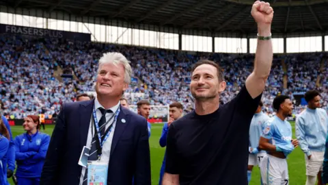 Coventry City Football Club purchases CBS Arena PA Media Coventry City owner Doug King pictured with manager Frank Lampard at the Coventry Building Society Arena celebrating with fans. They are both smiling and Frank Lampard has his left first raised in celebration.