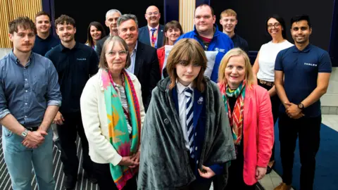 Pupil who invented device to help homeless named 'girl of the year' Thales A schoolgirl in school uniform, with a heated blanket wrapped around her. Several other people are standing around her smiling for the camera.
