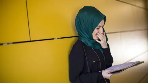GCSE grades 2025: The 9-1 boundaries explained Getty Images A young woman smiles as she looks at her GCSE results. She leans against a yellow wall and is wearing a dark jacket and a green headscarf.