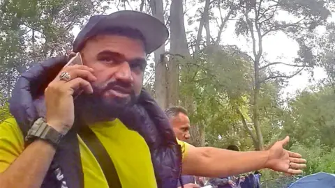 'Is my secret camera working?' Abdullah the smuggler, in a forest clearing, with people in the background behind him.