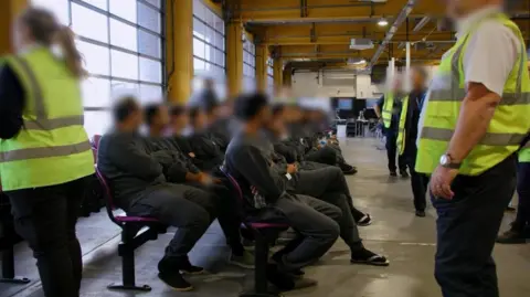 Government to overhaul asylum appeals system PA Media Staff in dark uniforms and hi-vis jackets look on as two rows of people in track suits and sandals sit waiting on two long rows of chairs in a large room that looks like a warehouse.