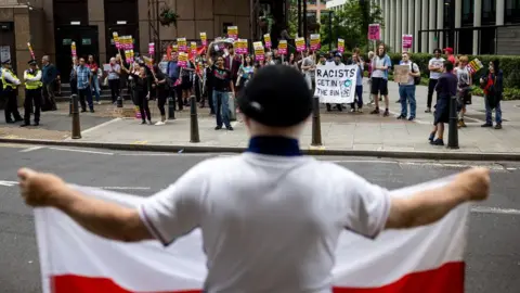 Behind the wave of asylum hotel protests Jack Taylor/Getty Images A man holds a flag bearing the St George's cross opposite protesters attending a rally organised by Stand Up To Racism outside the Britannia International Hotel on July 25, 2025 in London, England.