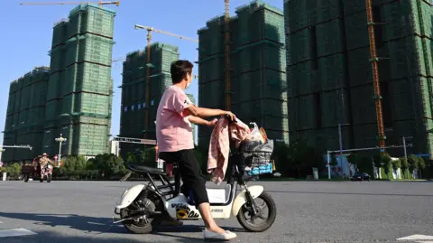 Chinese property giant to be delisted after spectacular fall AFP via Getty Images A woman - wearing a mask, pink t-shirt with a Mickey Mouse emblem on the right sleeve and black trousers - rides a scooter past the construction site of an Evergrande housing complex in Zhumadian, central China's Henan province.