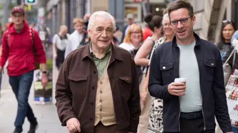 PA Media Actor Brian Cox  in green shirt and brown jacket walks along the Royal Mile in Edinburgh with James Graham, a bespectacled man in a blue jacket and green T shirt. He is carrying a disposable drinking cup.