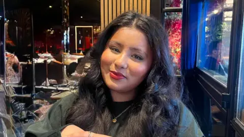 Working class students divided on civil service summer internship reform A photo of Hannah Begum sitting in a restaurant. She has long black hair and is wearing a black jumper with a golden pendant.