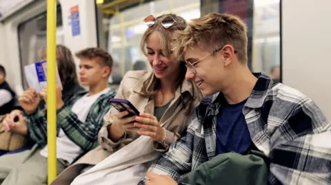 Transport for London (TfL) asks mobile users to wear headphones Getty Images A young adult male and a young adult female sit next to each other on a Tube looking at a mobile phone and smiling
