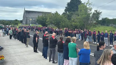 Victims of fatal shooting to be laid to rest in County Clare People lined up on a street outside a church. There is a cloudy sky above.