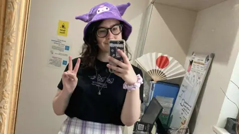 Claire's was glitz 'heaven' for kids before Shein and TikTok came along Al Thomann A person with shoulder-length wavy hair takes a selfie in a mirror, wearing a purple animal-shaped hat, a necklace and a black t-shirt. In the background is a computer and a fan with the Japanese flag
