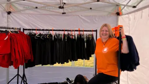 Forest of Dean school uniform pop-up a 'godsend', mum says Chippenham Uniform Exchange Jade Ugur is smiling and wearing a bright orange T-shirt. She is under a white gazebo with two rails of school uniform.