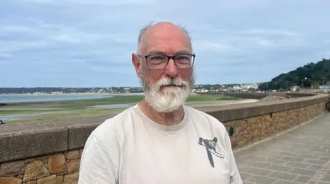 Falling birth rate in Jersey a 'ticking bomb' Nigel Jones is wearing a white T-shirt, he has a white beard and is wearing glasses.