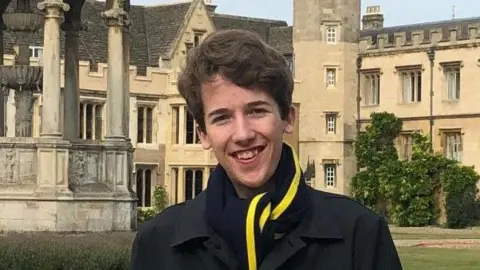 Working class students divided on civil service summer internship reform A photo of Peter Murphy standing in front of old university buildings. He is wearing a black coat and scarf with a yellow and black stripe. He has short brown hair.