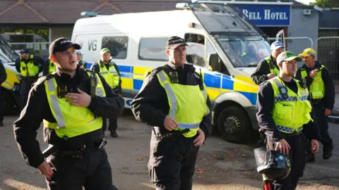 High court decision to be made on Epping asylum hotel injunction PA Media A group of police officers in fluorescent tabards speaking into talkback devices and holding riot helmets - there is a large police van in the background at the entrance to a building with a Bell Hotel sign