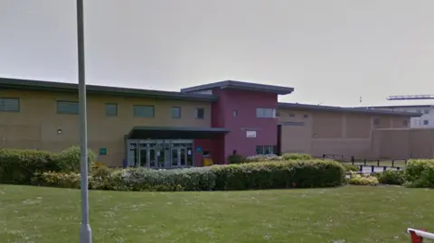 Lucy Connolly released from prison after race hate post on X Google Street view image of the prison from behind a barrier. It is a long rectangular building with a red on the walls where the entrance is. Outside is a stretch of grass with some bushes and flowers.