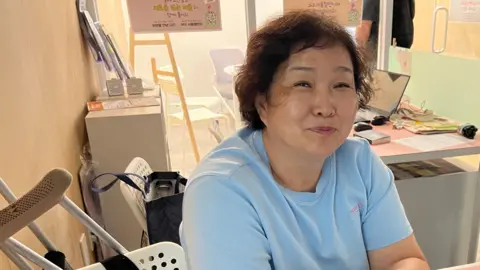 Jake Kwon/ BBC A smiling Lee In-sook at her desk in the store, in a blue t-shirt. 