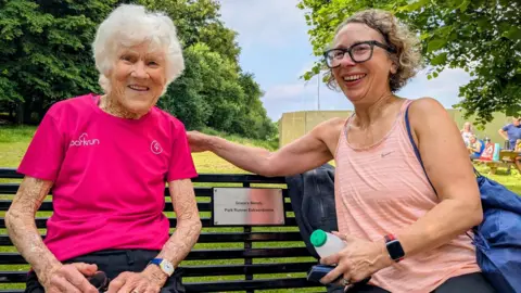 Parkrun record broken by 97-year-old Belfast runner Ormeau Parkrun Grace Chambers wearing a pink parkrun t-shirt. She is looking at the camera, She is sitting on a black park bench, there is a small plaque on the bench which says Grace's bench - Park Runner extraordinaire. She is sitting with another woman in a peach colour running top and black glasses