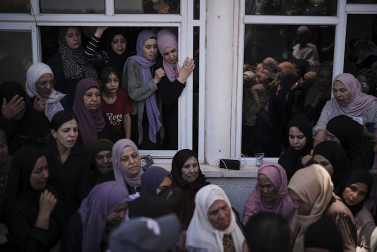 What’s fueling Israel’s surge of settler violence – and the lack of state response A crowd of women in dresses and headscarves look out of open windows.