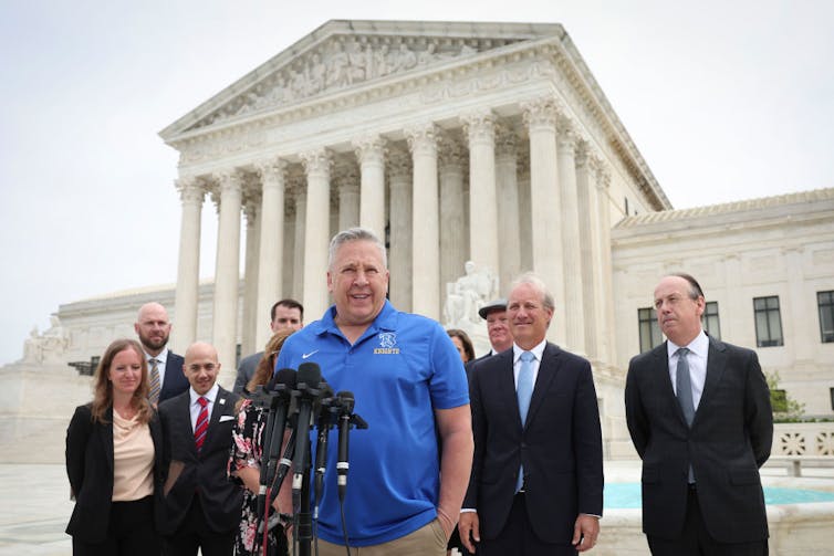 A man with silver hair who is wearing a short-sleeved blue polo stands before microphones, as a half-dozen people stand around him.