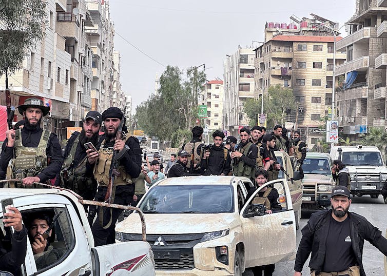 For Syrian Druze, latest violence is one more chapter in a centuries-long struggle over autonomy Two small white trucks with many soldiers standing on top of them drive through a wide city street.