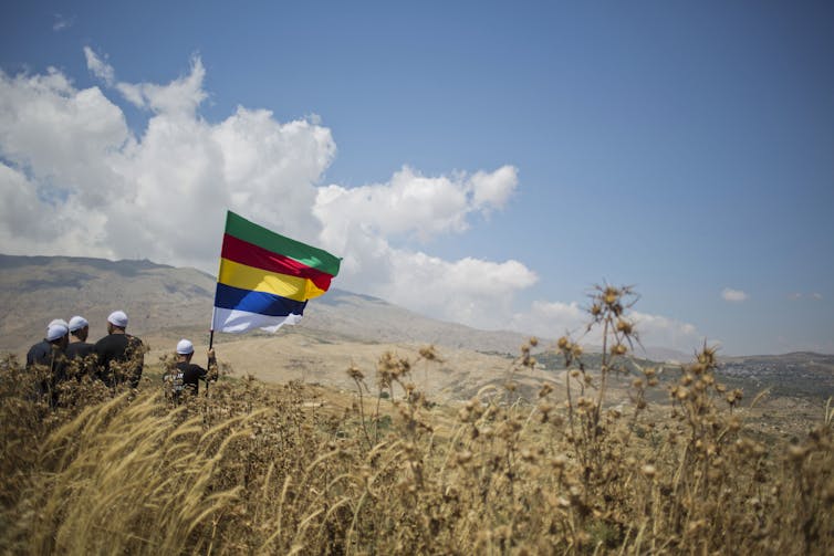 For Syrian Druze, latest violence is one more chapter in a centuries-long struggle over autonomy Four men in black clothing and white caps, seen from the back, hold a rainbow-striped flag as they stand in a vast field.