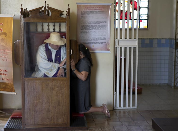 How Catholic confession has evolved over centuries A man in a white robe and straw hat sits inside a wooden cubicle as a woman keels beside it, with a small screen shielding her face.