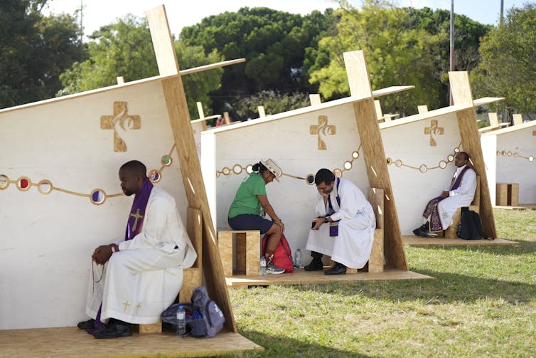How Catholic confession has evolved over centuries A row of open-air structures, with three walls for privacy, set up in a row, with a man in white robes sitting in each one.