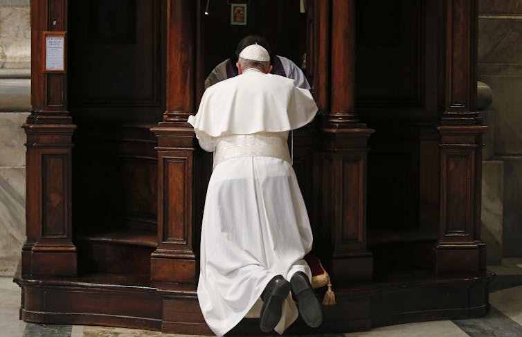 How Catholic confession has evolved over centuries A man in white robes and a white skullcap, seen from the back, kneeling in front of an ornate wooden cubicle.