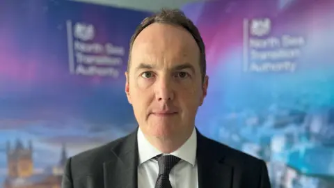 Net-zero 'not a platitude' for oil and gas sector BBC A man with brown hair wearing a dark tie, white shirt and dark suit stands in front of a blue wall with the North Sea Transition Authority's logos.