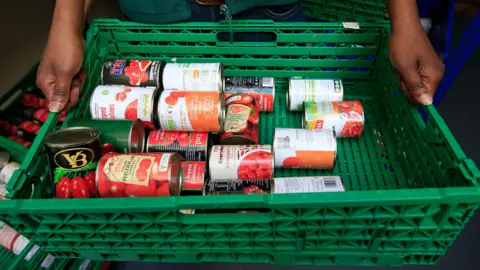 UK's first foodbank in Wiltshire celebrates 25th birthday PA Media A shot looking down on a plastic green crate full of various brands of tinned tomatoes being held by someone, though you can only see their hands.