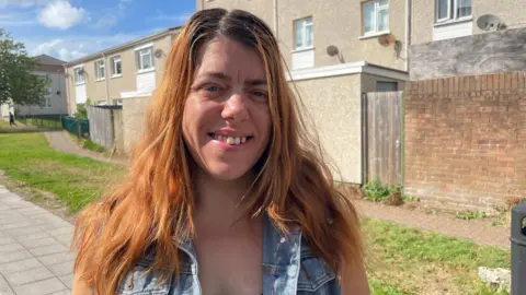 School costs a 'real struggle' for parents in Wales A woman with long auburn hair wearing a denim waistcoat standing in front of some terraced houses on an estate.