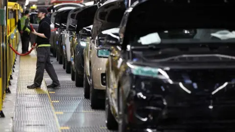 Reuters This generic image shows a member of staff on the left working on a car production line at Jaguar Land Rover's factory in Solihull. Cars are lined up with their bonnets up.
