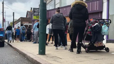 Birmingham parents debate costs in queue for school uniform A queue has formed outside a uniform shop. People are standing and waiting. Many are wearing jackets and carrying bags. One woman has a pushchair. A car is parked in the road. Other businesses can be seen in the street. There is a To Let sign on one property.