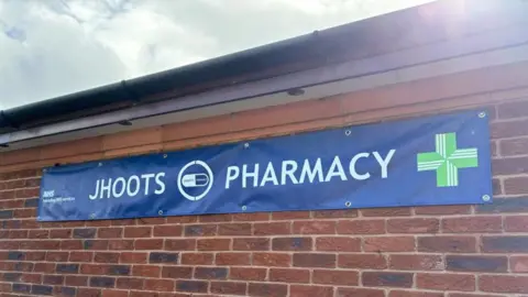 'Shocking' Jhoots pharmacy chain should be shut down, says MP BBC A Blue NHS branded banner which reads "Jhoots Pharmacy" is shown stuck to a brick wall. The NHS symbol can be seen in the far left, with the pharmacy symbol in the far right.