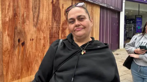 Birmingham parents debate costs in queue for school uniform Tracey Johnson is wearing a black top and gold necklace and she has sunglasses on her head. She is standing in the queue for the shop. Behind her, another business is boarded up. Another shopper, who is looking at her phone, can be seen behind her.