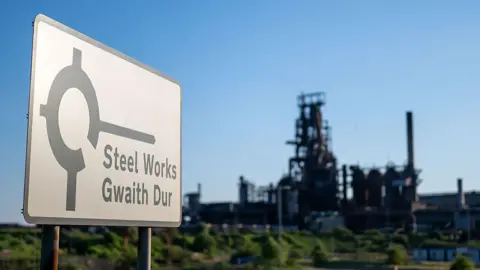 Getty Images A bilingual sign in the foreground reads "Steel Works / Gwaith Dur" beside an roundabout symbol. Behind it stands a large steelworks facility with tall chimneys and complex machinery, set against a clear blue sky. 